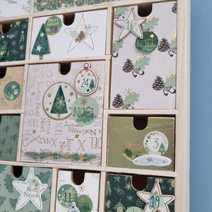Puede incluir: Un calendario de adviento de madera con m&uacute;ltiples cajones que presentan varios dise&ntilde;os navide&ntilde;os en tonos verde, blanco y dorado. Los cajones est&aacute;n decorados con ilustraciones de ciervos, estrellas, pi&ntilde;as y &aacute;rboles, junto con el texto Noel.