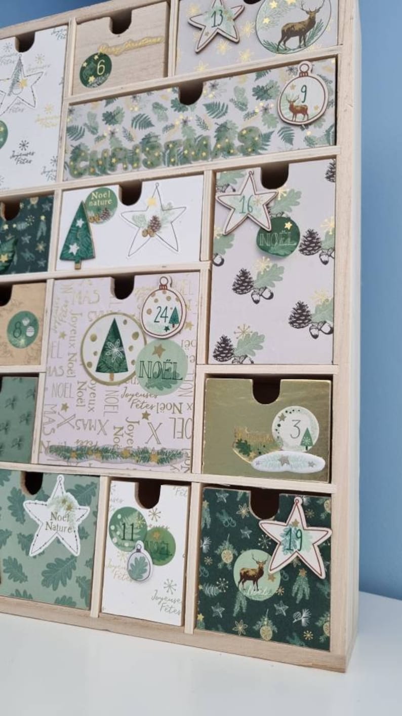 Puede incluir: Un calendario de adviento de madera con m&uacute;ltiples cajones que presentan varios dise&ntilde;os navide&ntilde;os en tonos verde, blanco y dorado. Los cajones est&aacute;n decorados con ilustraciones de ciervos, estrellas, pi&ntilde;as y &aacute;rboles, junto con el texto Noel.