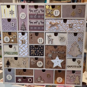 Puede incluir: Un calendario de adviento de madera con veinticuatro cajones individuales, cada uno con patrones, números y elementos decorativos únicos como estrellas, árboles, renos y copos de nieve. El cajón número 24 muestra el texto Joyeux Noel.