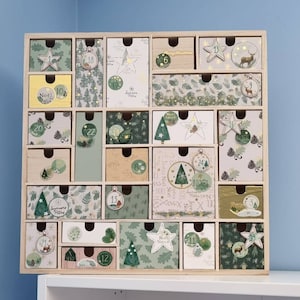 Puede incluir: Un calendario de adviento de madera cuadrado con veinticuatro cajones individuales. Cada caj&oacute;n presenta papel decorativo &uacute;nico en tonos verdes, blancos y beige, adornado con ilustraciones de pinos, estrellas, adornos y los textos No&euml;l, Christmas y Joyeuses F&ecirc;tes.
