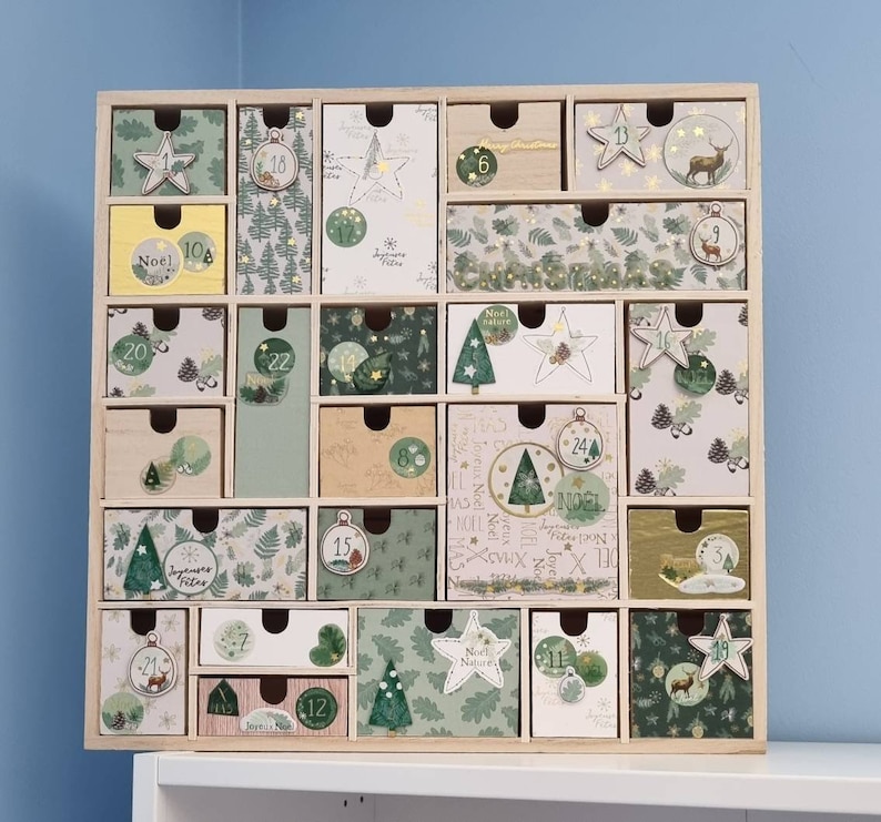 Puede incluir: Un calendario de adviento de madera cuadrado con veinticuatro cajones individuales. Cada caj&oacute;n presenta papel decorativo &uacute;nico en tonos verdes, blancos y beige, adornado con ilustraciones de pinos, estrellas, adornos y los textos No&euml;l, Christmas y Joyeuses F&ecirc;tes.