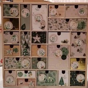 Puede incluir: Un calendario de adviento de madera con veinticuatro cajones individuales, cada uno con patrones de papel decorativos, ilustraciones festivas de &aacute;rboles, ciervos y pi&ntilde;as, y etiquetas circulares numeradas del uno al veinticuatro.