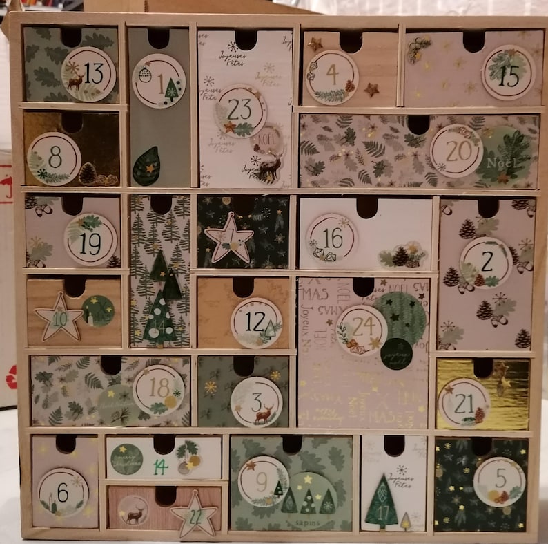 Puede incluir: Un calendario de adviento de madera con veinticuatro cajones individuales, cada uno con patrones de papel decorativos, ilustraciones festivas de &aacute;rboles, ciervos y pi&ntilde;as, y etiquetas circulares numeradas del uno al veinticuatro.