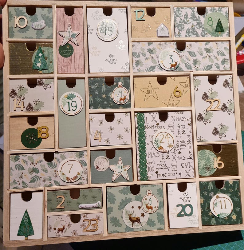 Puede incluir: Un calendario de adviento de madera con veinticuatro cajones individuales, cada uno decorado con papel tem&aacute;tico festivo en tonos verde, beige y blanco. Los cajones presentan n&uacute;meros del 1 al 24, junto con ilustraciones de ciervos, pinos y estrellas.