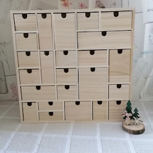 Puede incluir: Una unidad de almacenamiento de madera clara con veinticuatro cajones de distintos tama&ntilde;os, cada uno con un tirador circular recortado. A la derecha hay un peque&ntilde;o adorno decorativo de madera que representa un reno y dos pinos.