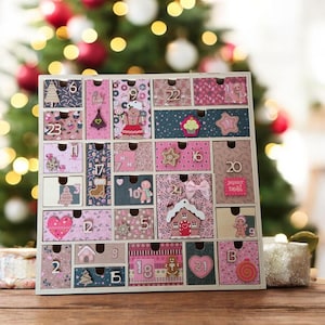 Puede incluir: Un calendario de adviento de madera con 24 cajones numerados. Cada cajón está decorado con motivos festivos, hombres de jengibre y adornos navideños. El calendario está situado frente a un fondo borroso de árbol de Navidad.
