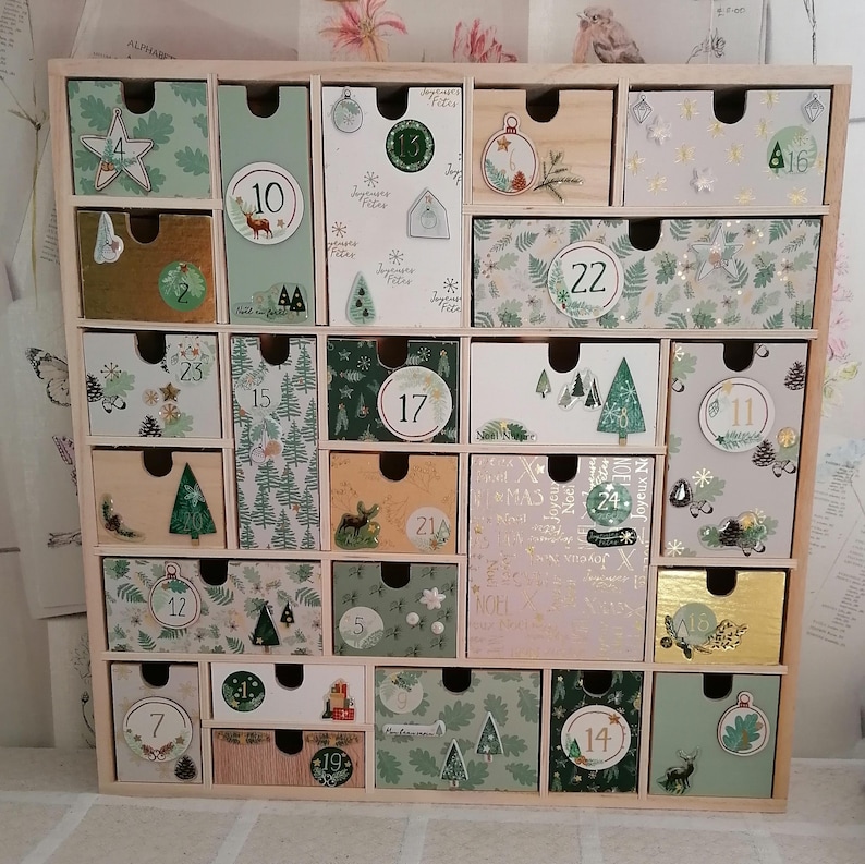 Puede incluir: Un calendario de adviento de madera con veinticuatro cajones individuales, cada uno con ilustraciones, patrones y n&uacute;meros tem&aacute;ticos festivos. Los cajones est&aacute;n dispuestos en una cuadr&iacute;cula dentro de un marco de madera clara, mostrando varios tonos de verde, blanco y dorado.