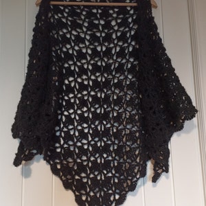 May include: A black crocheted shawl with a delicate openwork pattern. The shawl is draped over a wooden hanger.