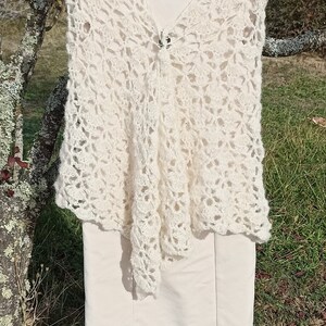 May include: A white crocheted vest with a delicate floral pattern. The vest is sleeveless and has a V-neckline. It is hanging on a hanger against a backdrop of green grass and a tree.