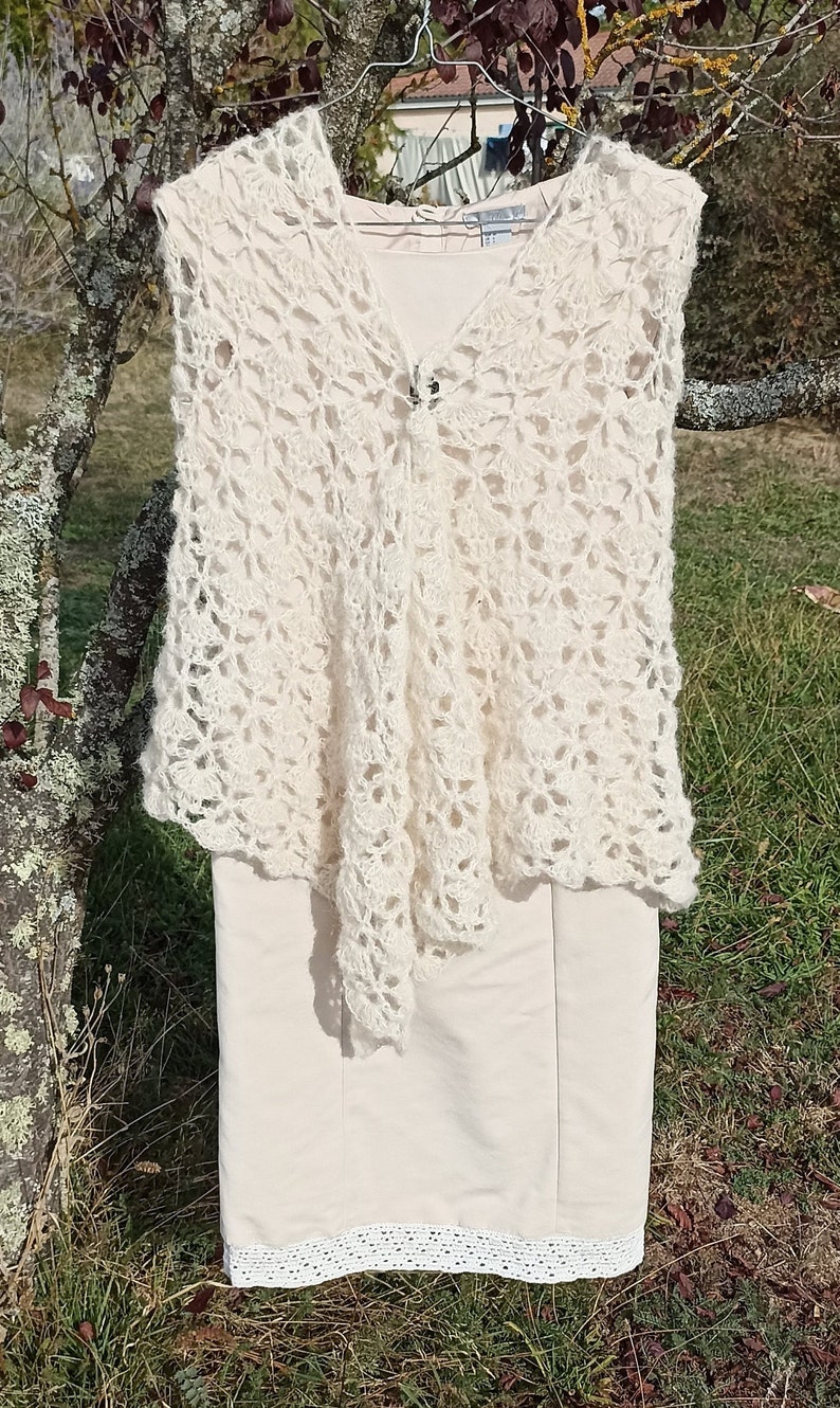 May include: A white crocheted vest with a delicate floral pattern. The vest is sleeveless and has a V-neckline. It is hanging on a hanger against a backdrop of green grass and a tree.