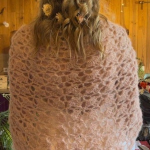 May include: A light pink crocheted shawl with a delicate floral pattern. The shawl is draped over the shoulders of a person with long, curly hair adorned with a floral crown.