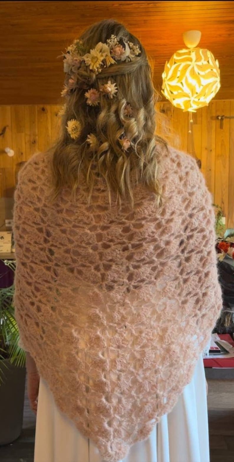 May include: A light pink crocheted shawl with a delicate floral pattern. The shawl is draped over the shoulders of a person with long, curly hair adorned with a floral crown.