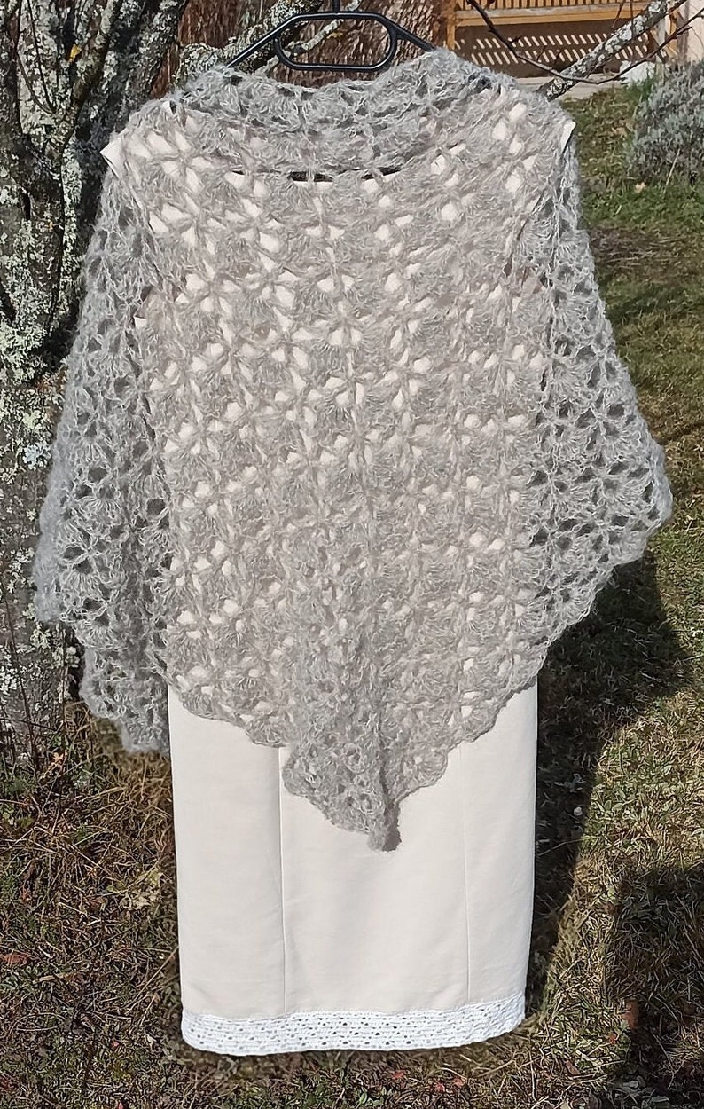 May include: A gray crocheted shawl with a delicate openwork pattern. The shawl is draped over a white dress with a lace trim.