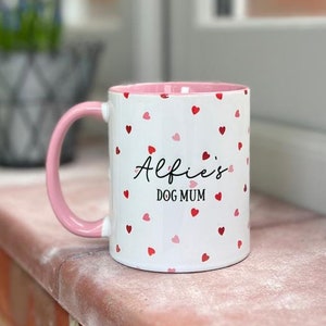 May include: White ceramic mug with a pink handle and interior, decorated with red and pink hearts. The mug features the text "Alfie's DOG MUM" in black script. A charming gift for dog lovers.