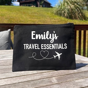 May include: Black canvas travel makeup bag with white text that reads "Emily's Travel Essentials" and a white airplane with a heart above it.