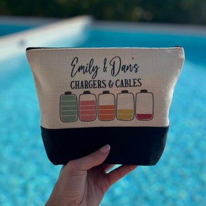 May include: A beige canvas makeup bag with black trim. The bag has the text "Emily & Dan's Chargers & Cables" printed on it. Five colorful battery icons are below the text.