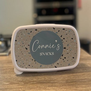 May include: White rectangular lunchbox with a terrazzo pattern and a blue circle with the text "Connie's Snacks" in white.
