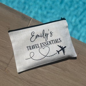 May include: A white canvas zippered pouch with black text that reads "Emily's Travel Essentials" and a black airplane with a heart-shaped trail.