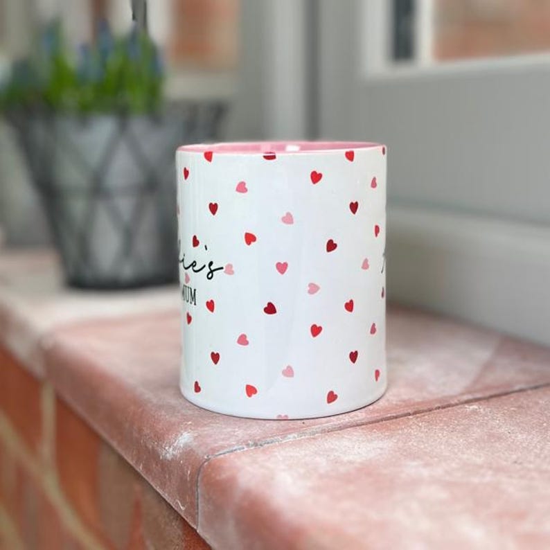 May include: White ceramic mug with a pink interior, covered in red and pink heart shapes. The mug has the text "ie's Mum" in black script. The mug is sitting on a brick ledge.