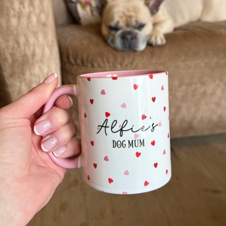 May include: White ceramic mug with pink handle and interior, decorated with red and pink hearts. The mug has the text "Alfie's DOG MUM" in black script. A dog is resting in the background.