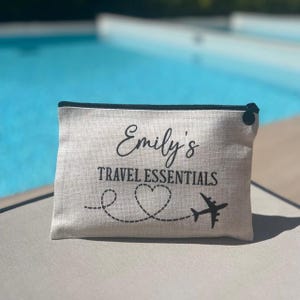 May include: A white canvas travel pouch with a black zipper and black trim. The pouch has the text "Emily's Travel Essentials" printed on it, along with a heart and a plane.