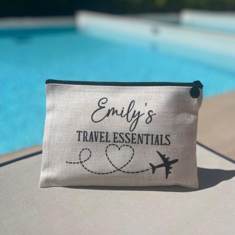 May include: A white canvas travel pouch with a black zipper and black trim. The pouch has the text "Emily's Travel Essentials" printed on it, along with a heart and a plane.