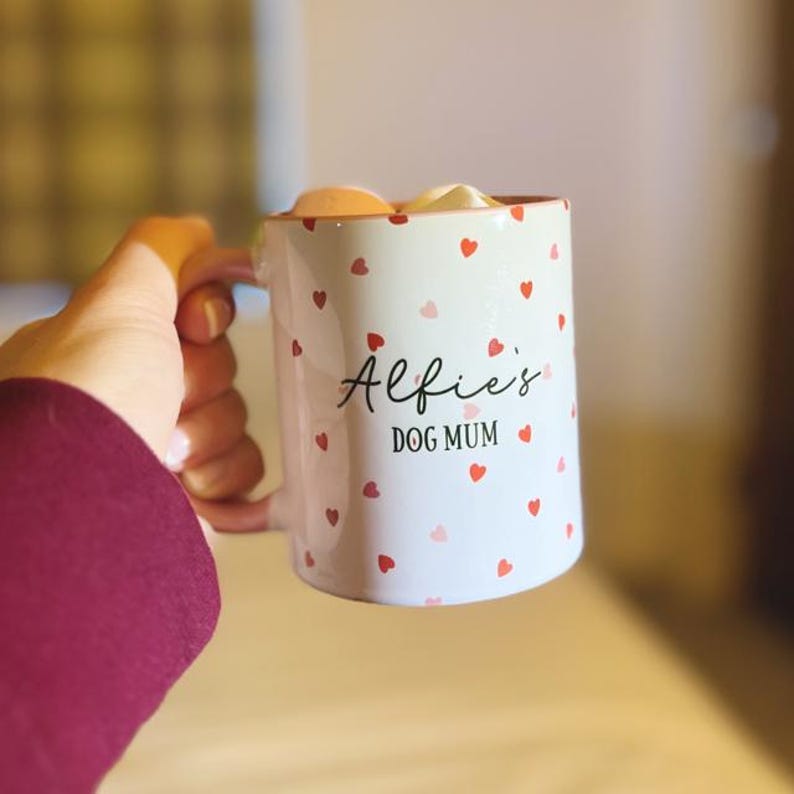May include: White ceramic mug decorated with red and pink hearts. The mug has the text "Alfie's DOG MUM" in black script. The mug is filled with marshmallows. A hand is holding the mug.