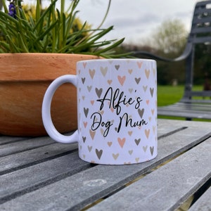 Puede incluir: Taza de cer&aacute;mica blanca con un patr&oacute;n de corazones y el texto "Alfie's Dog Mum" en escritura negra.