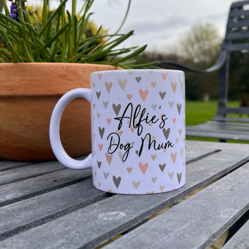 Puede incluir: Taza de cer&aacute;mica blanca con un patr&oacute;n de corazones y el texto "Alfie's Dog Mum" en escritura negra.