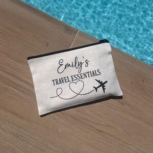 May include: A white canvas travel makeup bag with black trim and the text "Emily's Travel Essentials" with a heart and airplane graphic.