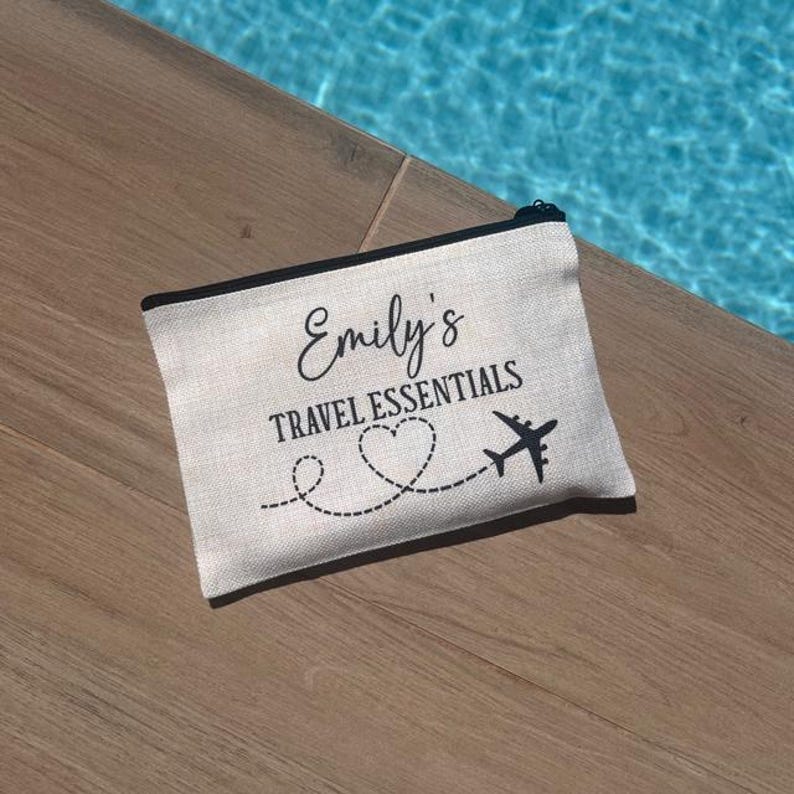 May include: A white canvas travel makeup bag with black trim and the text "Emily's Travel Essentials" with a heart and airplane graphic.