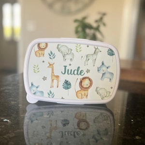 May include: White lunchbox with a jungle animal print design. The lunchbox has a green name tag that says "Jude".