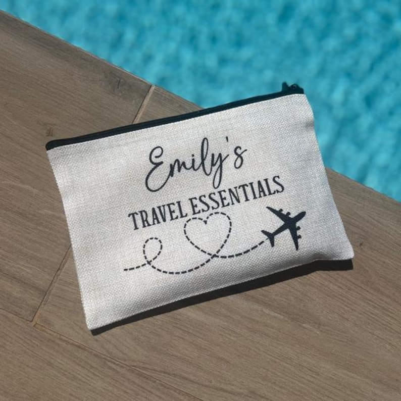 May include: A white canvas zippered pouch with black text that reads "Emily's Travel Essentials" and a black airplane with a heart-shaped trail.