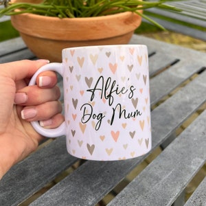 Puede incluir: Taza de cer&aacute;mica blanca con un patr&oacute;n de corazones rosas y marrones. La taza tiene un texto negro que dice "Alfie's Dog Mum".