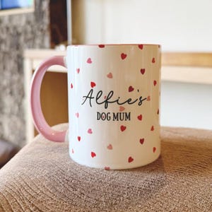 May include: White ceramic mug with a pink handle and rim, decorated with red and pink hearts. The mug features the text "Alfie's DOG MUM" in black script. A charming and personalized gift for dog lovers.