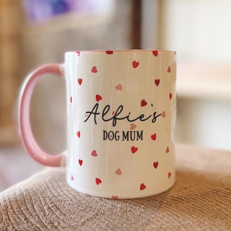 May include: White ceramic mug with a pink handle and rim, decorated with red and pink hearts. The mug features the text "Alfie's DOG MUM" in black script, making it a perfect gift for a dog lover.