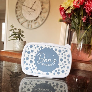 May include: A white lunchbox with a blue lid featuring a white polka dot pattern. The lid has a blue circle with the text "Dan's Snacks" in white cursive writing.