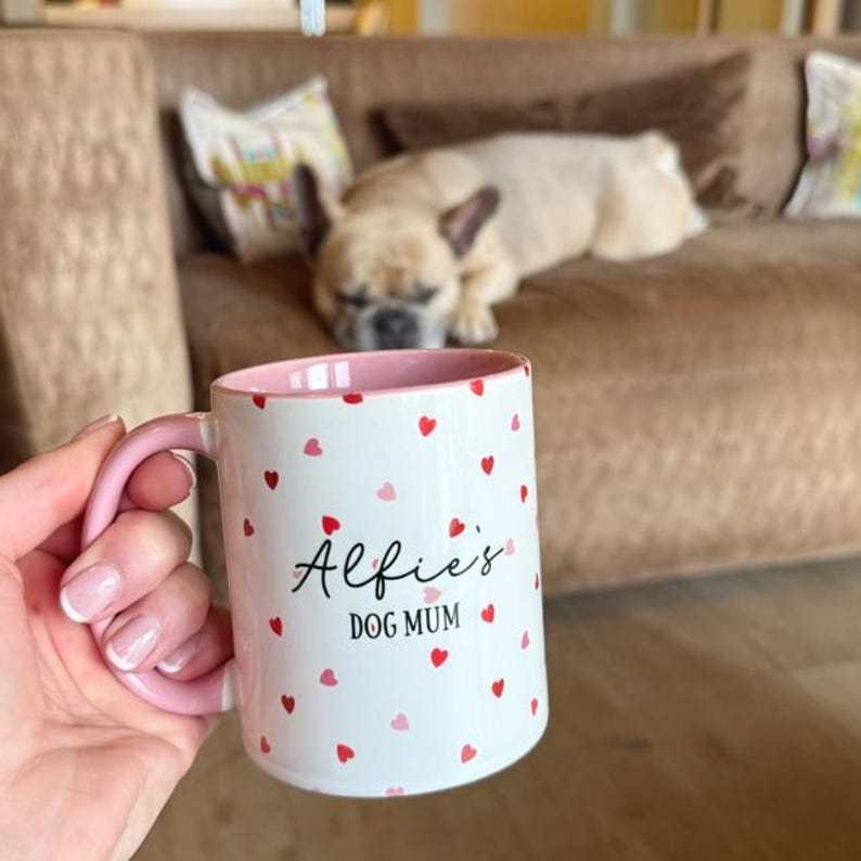 May include: White ceramic mug with a pink handle and interior, decorated with red and pink hearts. The mug reads "Alfie's DOG MUM" in black script. A dog is resting on a brown couch in the background.
