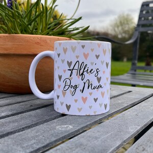 Puede incluir: Taza de cer&aacute;mica blanca con un patr&oacute;n de corazones en tonos rosa y marr&oacute;n. La taza tiene el texto "Alfie's Dog Mum" en negro.