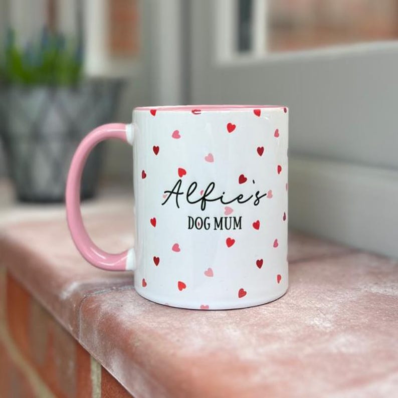 May include: White ceramic mug with a pink handle and rim, decorated with red and pink hearts. The mug has the text "Alfie's DOG MUM" in black script. A charming gift for a dog owner.