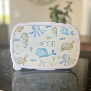 May include: White lunchbox featuring a blue and yellow sea creature design. The lunchbox has the name "JACOB" printed on the front.