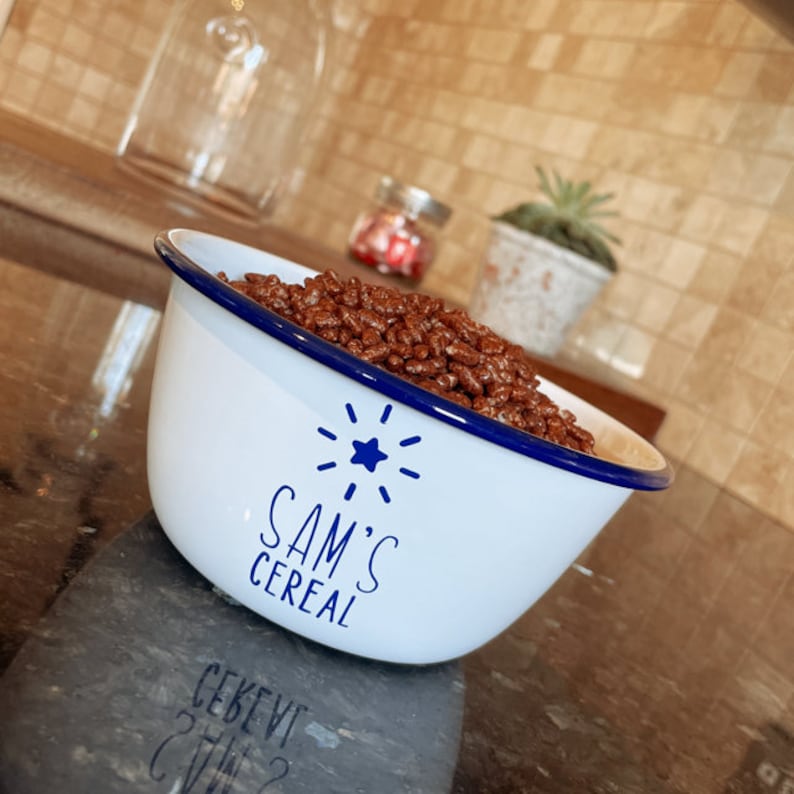 May include: A white enamel bowl with a blue rim and a blue starburst design. The bowl says "Sam's Cereal" and is filled with chocolate cereal.
