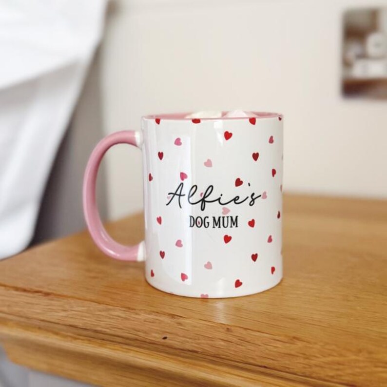 May include: White ceramic mug with a pink handle and rim, adorned with red and pink heart patterns. The mug features the text "Alfie's DOG MUM" in a script font, perfect for dog lovers. The mug is sitting on a wooden surface.