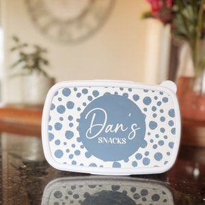 May include: White lunchbox with a blue and white polka dot pattern. The lunchbox has a blue circle with the text "Dan's Snacks" in white.