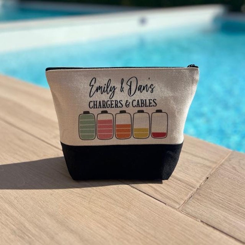 May include: A beige canvas zippered pouch with black trim. The pouch has the text "Emily & Dan's Chargers & Cables" printed on it. There are five battery icons with different colored levels printed on the pouch.