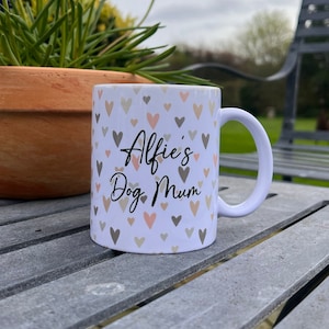Puede incluir: Taza de cer&aacute;mica blanca con un patr&oacute;n de corazones en tonos de rosa y gris. La taza tiene el texto "Alfie's Dog Mum" en negro.