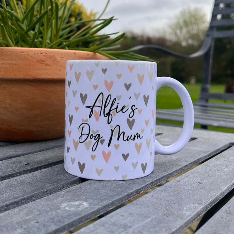 Puede incluir: Taza de cer&aacute;mica blanca con un patr&oacute;n de corazones en tonos de rosa y gris. La taza tiene el texto "Alfie's Dog Mum" en negro.
