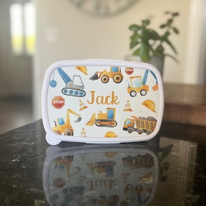 May include: White lunchbox with a construction theme. The lunchbox has a white background with yellow construction vehicles, orange traffic cones, and yellow hard hats. The name "Jack" is printed in orange on the front of the lunchbox.