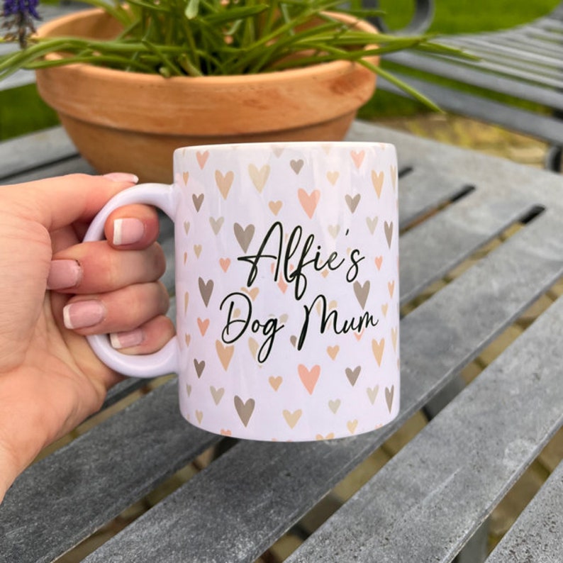 Puede incluir: Taza de cer&aacute;mica blanca con un patr&oacute;n de corazones en tonos marrones y rosas. La taza tiene el texto "Alfie's Dog Mum" en negro.
