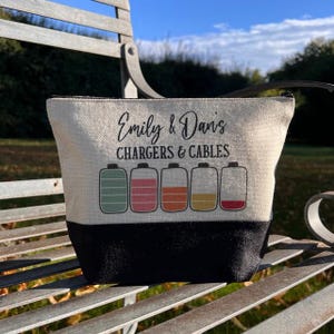 May include: A white canvas zippered pouch with black trim. The pouch has the text "Emily & Dan's Chargers & Cables" printed on the front. Five colorful battery icons are printed below the text.